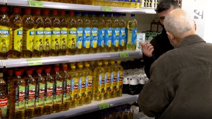 Dos personas dialogan frente a una fila de botellas de aceite.