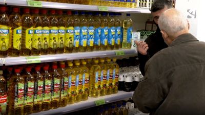 Dos personas dialogan frente a una fila de botellas de aceite.